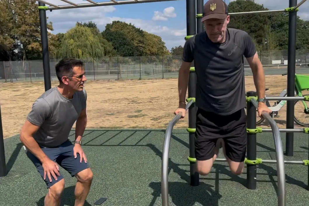 Linz crouching to watch a client hold a dip on parallel bars in an outdoor callisthenics park.