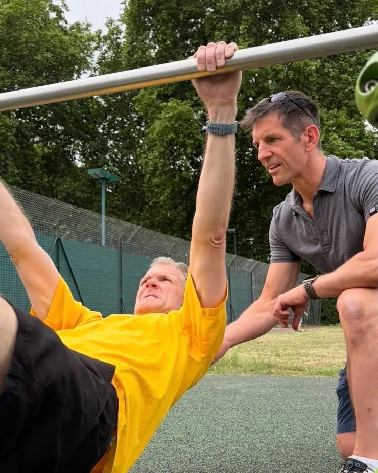Linz crouches beside a man in a yellow shirt who is hanging beneath an outdoor bar in a park, body held in a straight line, while Linz guides his shoulder.