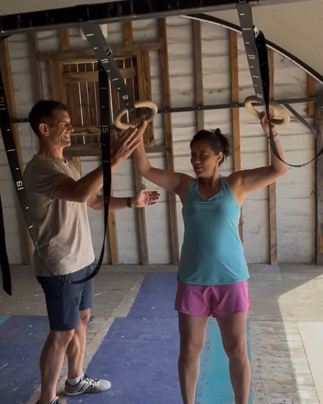 A woman holding a pair of gymnastic rings at shoulder height in a timber-walled home gym, smiling, while Linz stands beside her guiding the movement with his hands.