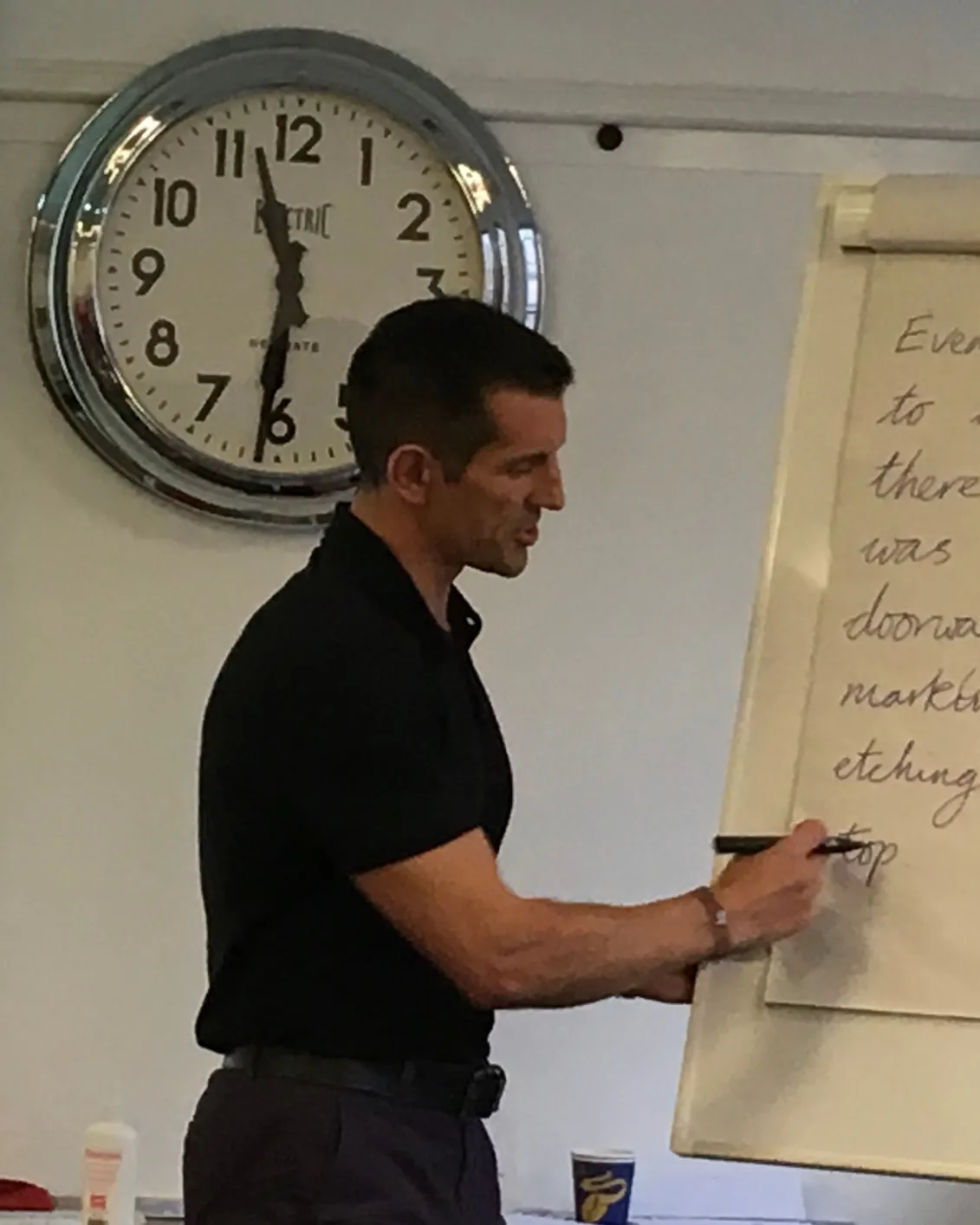 Linz writing on a flipchart at the front of a training room, a large wall clock behind him.